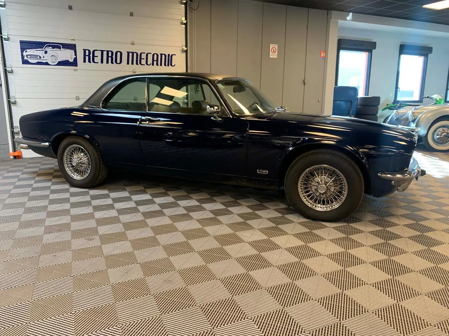 Jaguar XJ coupé bleu nuit à roues à rayons, devant le logo Retro Mecanic