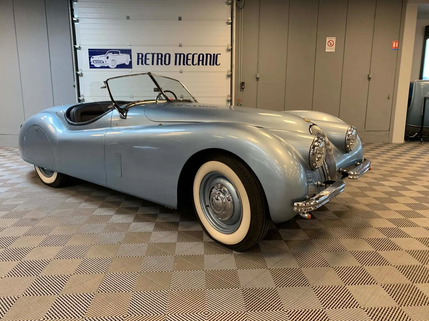 Jaguar XK120 roadster bleu argenté à pneus à flancs blancs, devant le logo Retro Mecanic